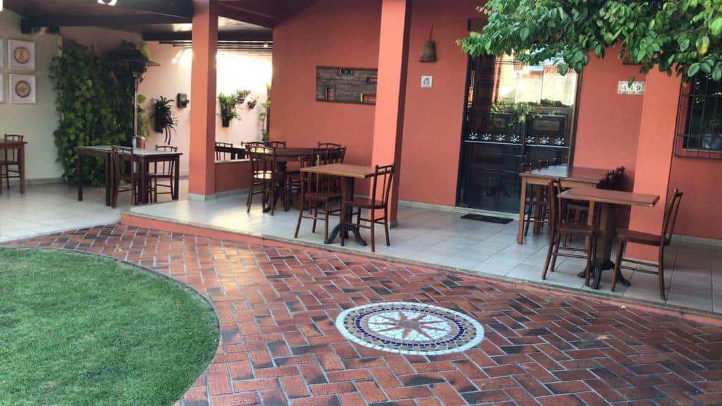 Parte da Pousada Bartrô com algumas mesas e cadeiras de madeira espalhadas na parte coberta da construção e algumas plantas em volta, ilustrando post Pousadas em Rio das Ostras.