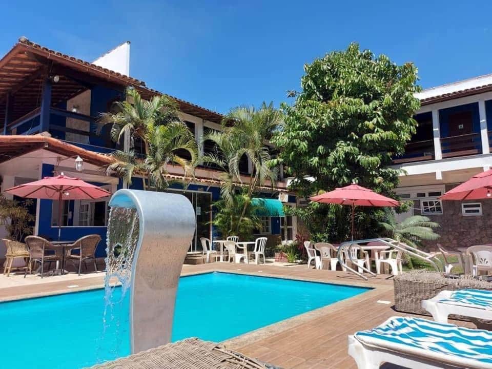 Parte da Pousada Belmare Eireli que mostra a piscina azul com uma cascata e em volta alguma espreguiçadeiras, cadeiras e mesas com guarda-sóis. Ao fundo tem algumas árvores verdes e a construção de dois andares da pousada durante o dia, ilustrando post Pousadas em Rio das Ostras.