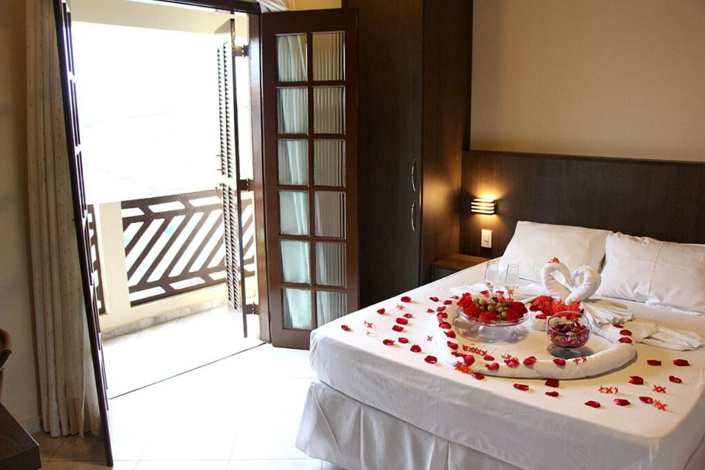 Quarto da Pousada Berro D'água. Uma cama de casal está ao lado direito, toda coberta por pétalas de rosa, frutas, doces e outras decorações românticas. Do lado esquerdo está a porta para a varanda.