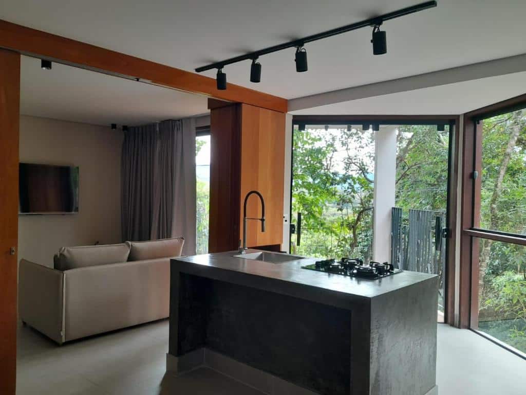 Imagem da Pousada Canto da Lua, vemos uma cozinha integrada com a sala. No meio, há uma bancada de mármore com a pia e um cooktop. Na frente, há uma grande porta de vidro e janelas, que dão vista para várias árvores. Dentro do ambiente, mais para o lado esquerdo da foto, vemos o sofá e costas e um TV n sua frente, fixa na parede.