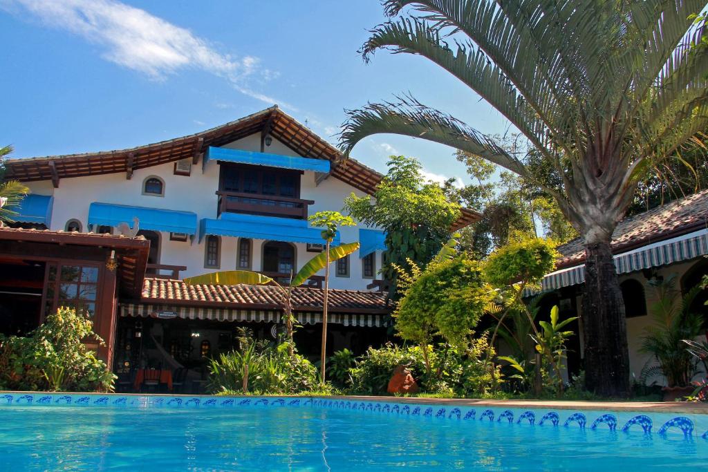 Parte da Pousada do Marujo que mostra a piscina azul com várias árvores em volta e ao fundo a construção da pousada com três andares durante o dia, ilustrando post Pousadas em Rio das Ostras.