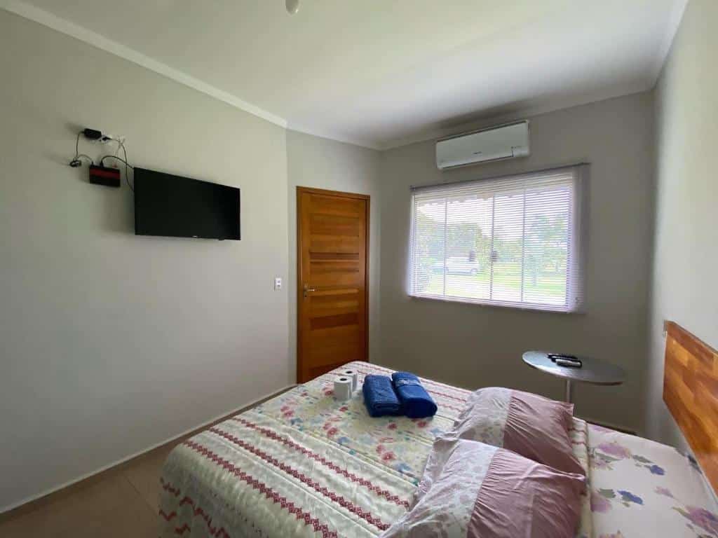 Quarto da Pousada Ilha Sul. Uma cama de casal está no meio, com a cabeceira encostada na parede. Em frente e pendurada na parede está a tv e, ao seu lado, a porta. Na parede ao lado da cama há uma janela e uma mesinha redonda.