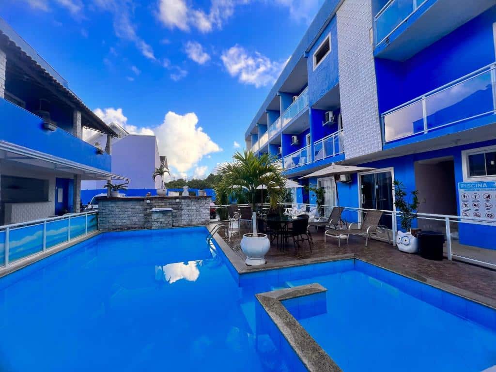 Parte da Pousada Maresia Costa Azul que mostra a piscina com uma parte funda e a a direita uma parte mais rasa. Em volta tem algumas cadeiras, guarda-sóis e plantas e fora do espaço da piscina tem a construção de três andares da pousada em azul durante o dia, ilustrando post Pousadas em Rio das Ostras.
