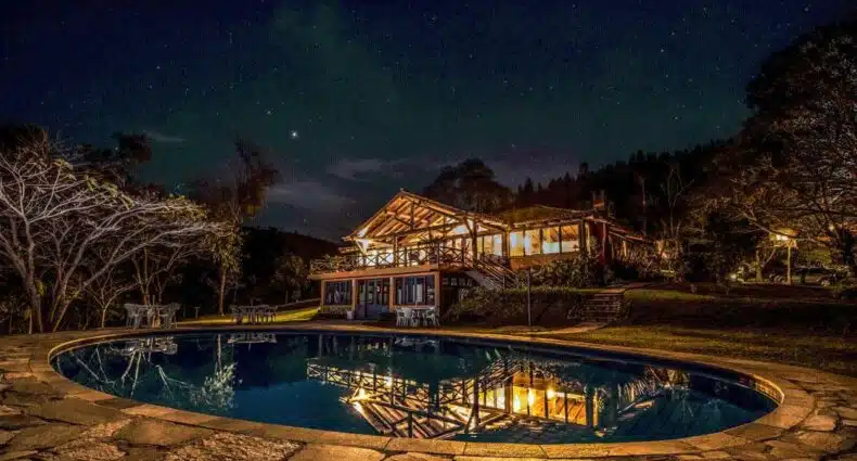 Área da piscina da Pousada Maria Manhã. A piscina tem um formato mais oval e está perto de uma região com grama e árvores. Ao fundo é possível a própria pousada. Imagem para o post de pousadas em Passa Quatro.