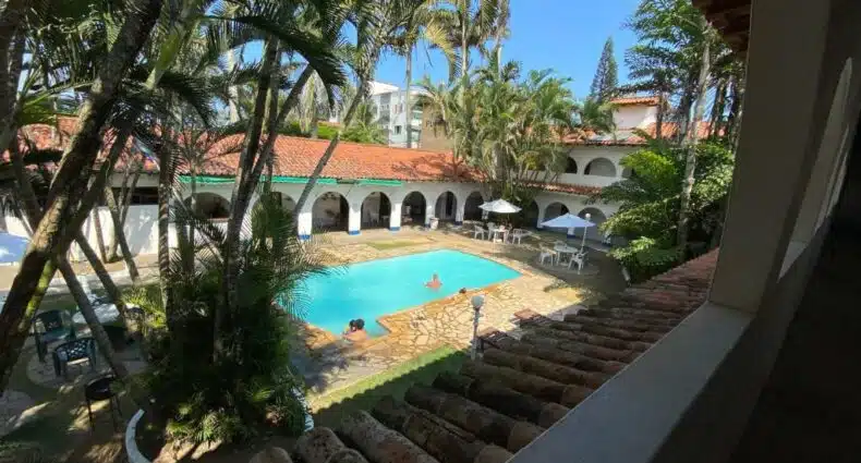 Uma piscina no pátio da Pousada Puerta del Sol, cercada por palmeiras e edifícios em arco, com pessoas nadando e sentadas em mesas, para representar pousadas em Rio das Ostras.