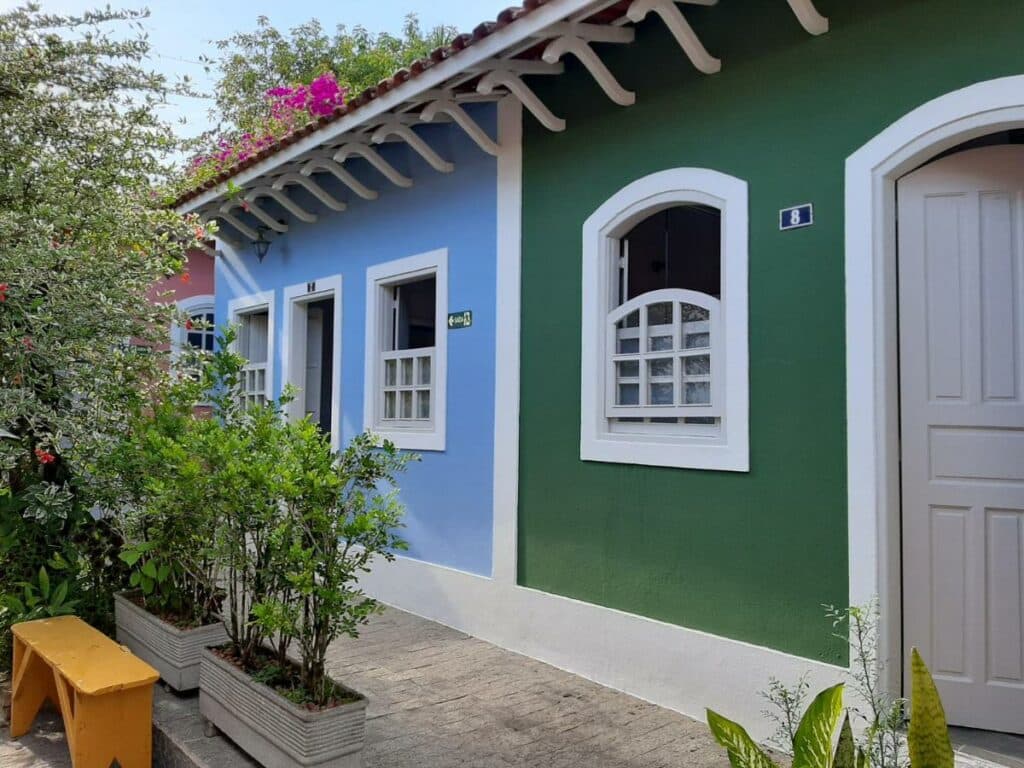 Lado de fora dos quartos da acomodação. Eles possuem portas estreitas e janelas de tamanho médio. Em frente existem várias plantas e flores, para representar pousadas em Cananéia.