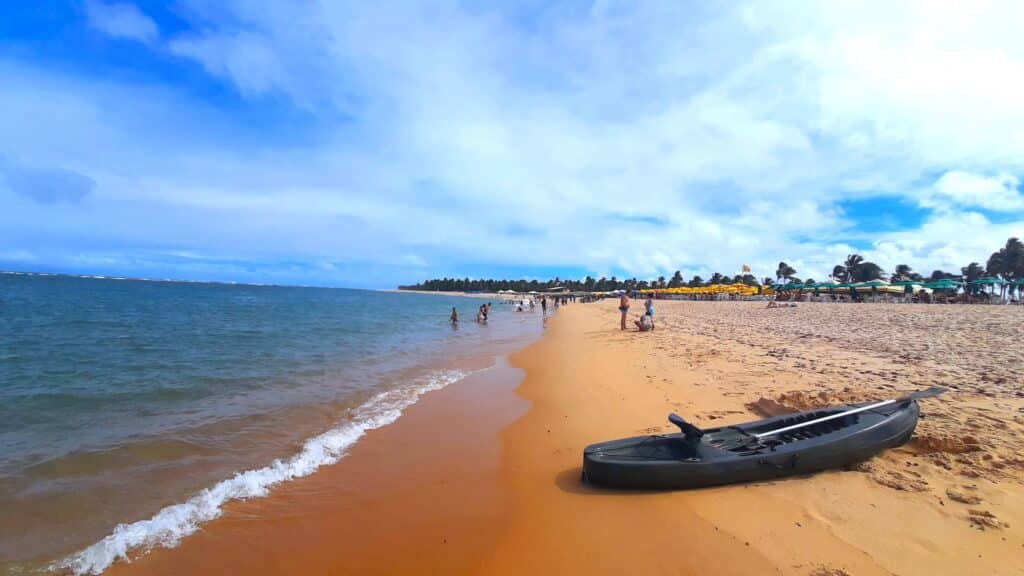 Praia do Gunga, em Alagoas, com um caiaque na reia, algumas pessoas nadando e de pé na areia e as mesas espalhadas dos quiosques