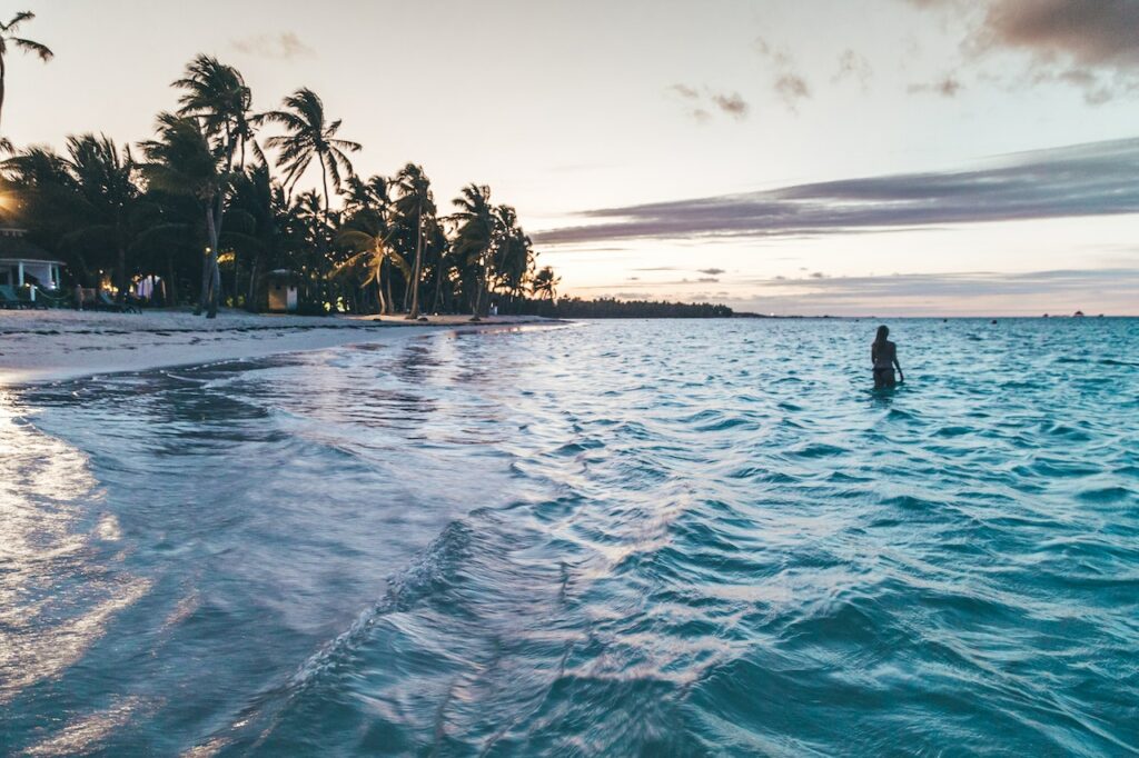 Foto tirada dentro do mar de uma mulher andando ao longe. No canto superior esquerdo há várias palmeiras. - Foto: Leonardo Rossatti via Pexels