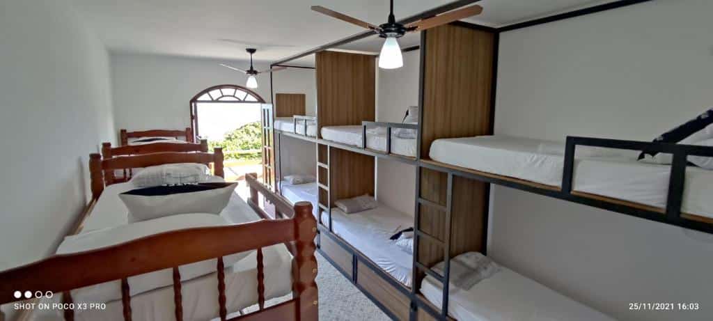 Quarto do Praia Virgem Hostel com cinco beliches de solteiro e um espaço livre no meio deles para andar, no teto tem dois ventiladores e ao fundo tem uma porta de vidro aberta, ilustrando post Pousadas em Rio das Ostras.