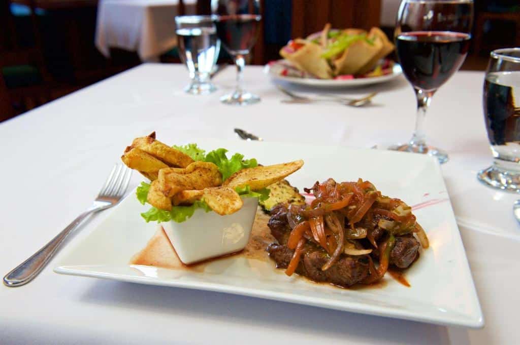 Foto de uma refeição no Radisson Fort George Hotel & Marina. A imagem foca em um prato com carne e acompanhamentos. A toalha da mesa é branca e no fundo vemos algumas taças com vinho e outro prato do outro lado da mesa.