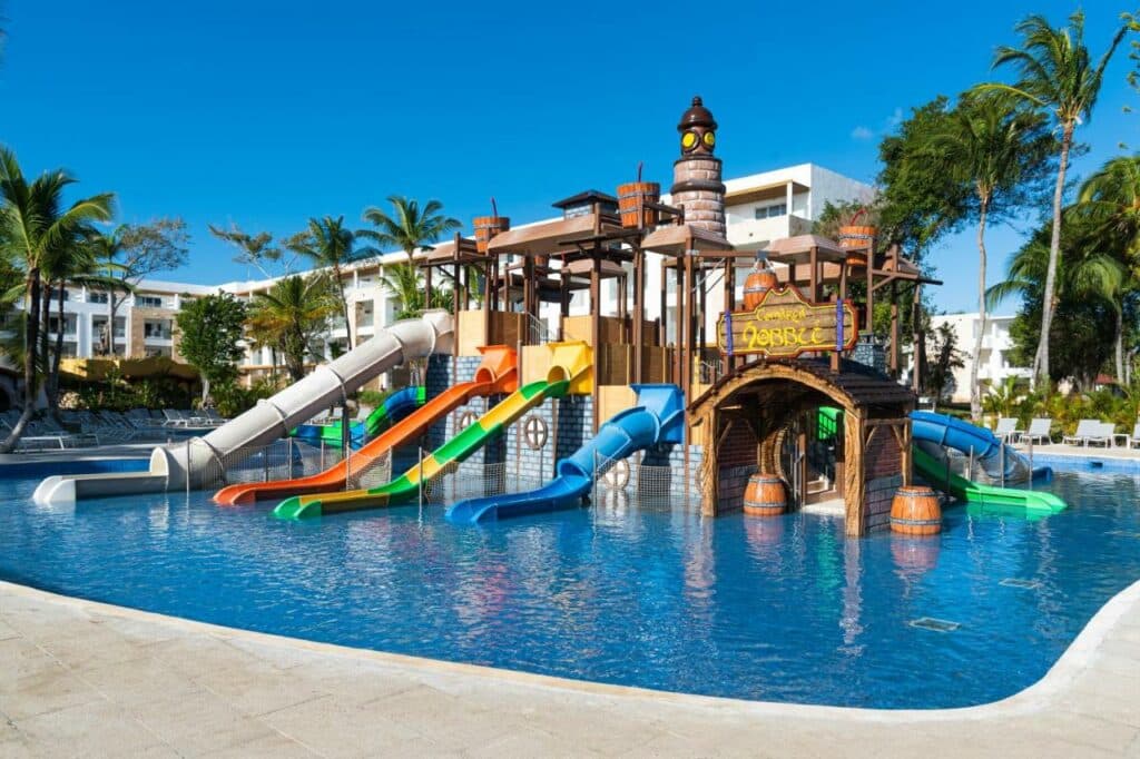 Parque aquático do Princess Family Club Bavaro com vários tobogãs e brinquedos no centro de uma piscina. Várias palmeiras rodeiam o local.