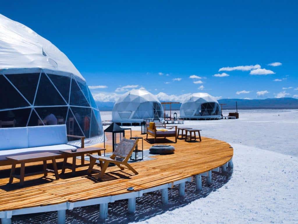 No lado esquerdo uma cabana do Pristine Camps na frente e duas atrás. Um deck na cabana com cadeiras de tomar sol e banquinhos com almofadas. Foto para ilustrar post sobre hotéis em Jujuy.