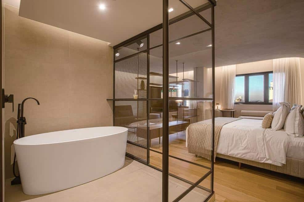 Quarto do QOYA Hotel Curitiba. Uma cama de casal do lado direito, no fundo uma televisão de frente. Do lado esquerdo a banheira do quarto com uma parede de vidro. Foto para ilustrar post sobre hotéis em Curitiba.