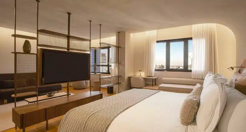 Quarto do QOYA Hotel Curitiba, Curio Collection by Hilton. Uma cama de casal do lado direito com um sofá no fundo. Do lado esquerdo um painel no meio do quarto com uma televisão. No fundo a janela do quarto. Foto para ilustrar post sobre hotéis em Curitiba.