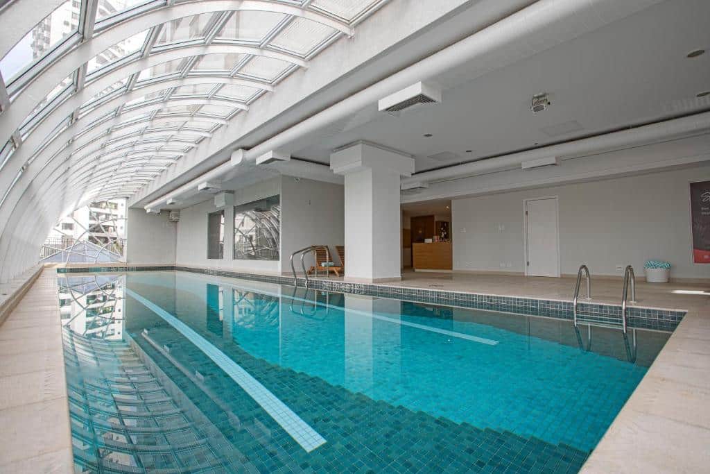 Piscina do QOYA Hotel Curitiba. Uma piscina coberta no meio, e do lado esquerdo parede e teto de vidro.