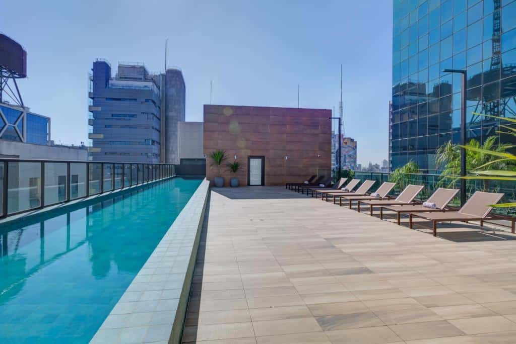 Piscina no terraço do Qoya São Paulo Paulista. Uma piscina no lado esquerdo, com uma parede de vidro. No lado direito cadeiras de tomar sol. Ao redor prédios.