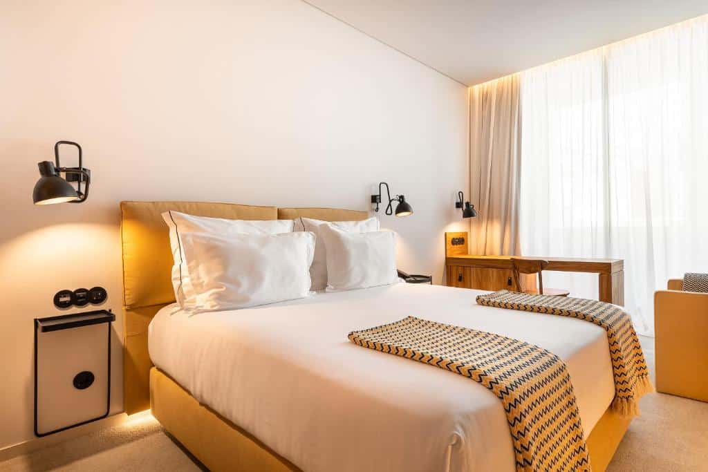 Quarto do 3HB Faro com cama de casal do lado esquerdo da imagem no centro, do lado esquerdo da imagem uma mesa de trabalho com cadeira. Representa hotéis para lua de mel em Portugal.