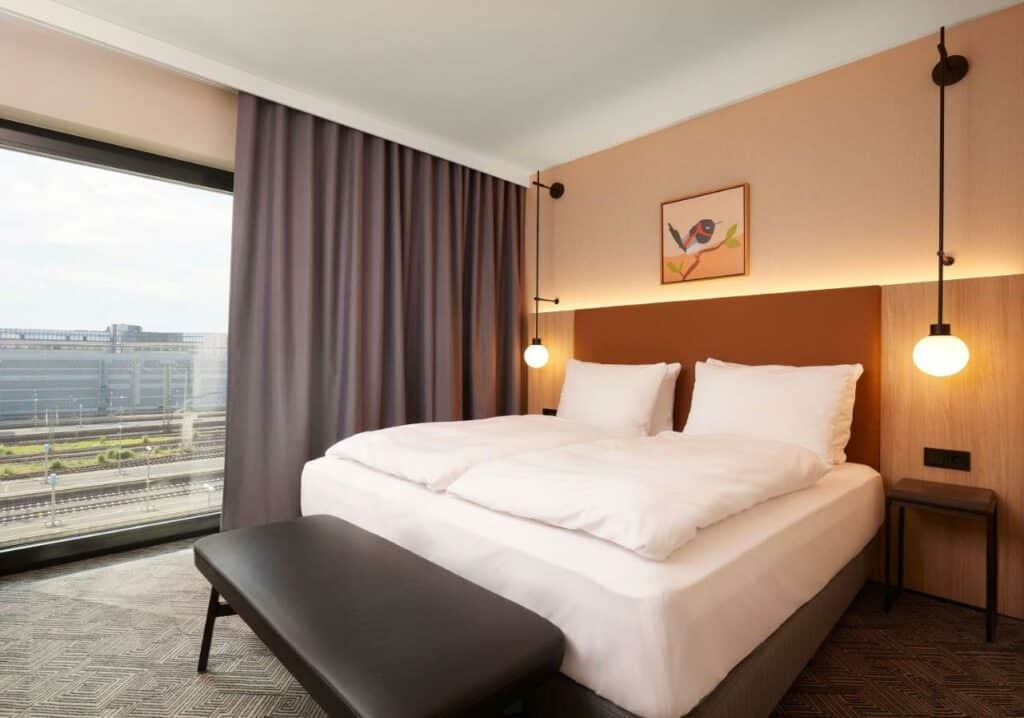 Quarto do Adina Apartment Hotel Dusseldorf com cama de casal do lado direito da imagem com banco ao pé da cama do lado esquerdo da cama janelas panorâmicas com vista para a cidade. Representa hotéis em Dusseldorf.