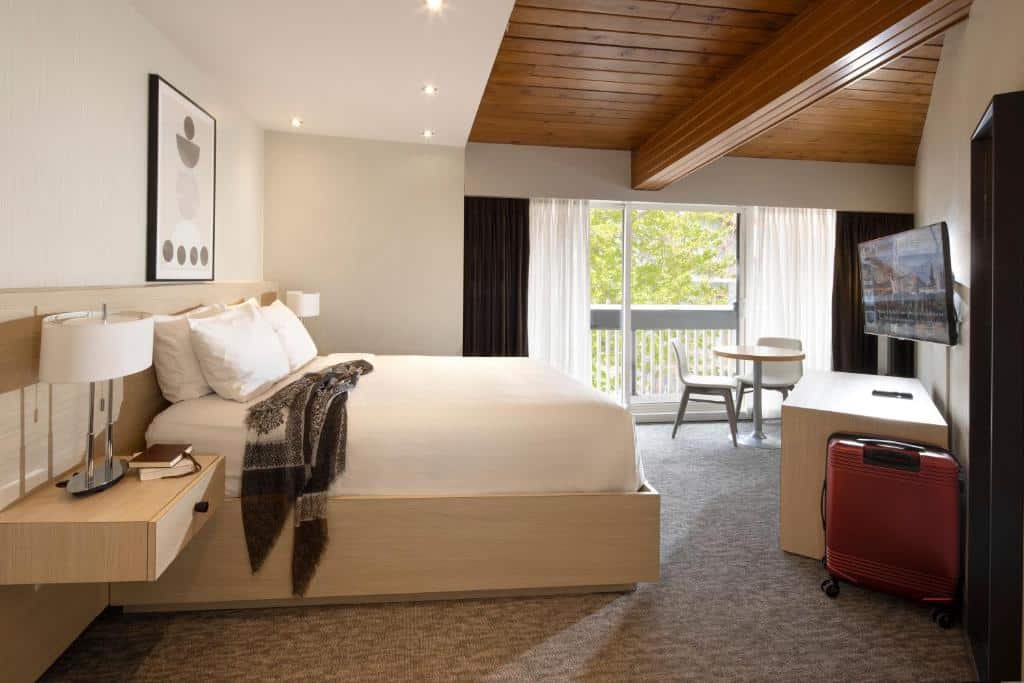Quarto do Banff Aspen Lodge com uma cama de casal do lado uma mesinha com um abajur e em cima na parede um quadro. O chão é de carpete, tem uma porta de vidro que dá acesso a sacada, há também uma mesinha redonda com duas cadeiras e em frente a cama tem uma mesa com uma tv na parede e uma mala.