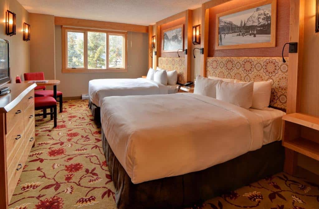 Quarto do Banff Ptarmigan Inn com duas camas de casal com travesseiros em cima e cobertas e de cada lado da cama tem uma mesinha de madeira. O chão é de carpete florido, tem dois quadros na parede, uma janela de vidro, duas cadeira vermelhas e uma mesa e ao lado uma cômoda com uma tv.