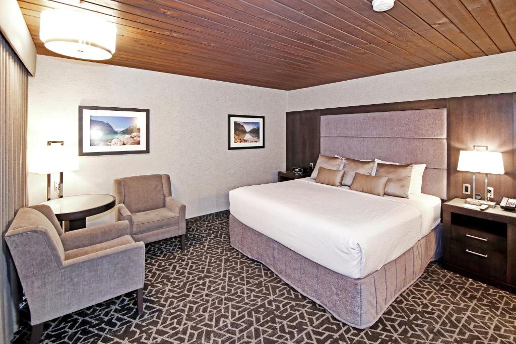 Quarto do Best Western Plus Siding 29 Lodge com uma cama de casal com várias almofadas em cima, cada lado da cama tem uma mesinha de madeira e uma delas tem um abajur. O chão é de carpete e o teto de madeira, há dois quadros na parede e uma mesa redonda com duas poltronas em volta.