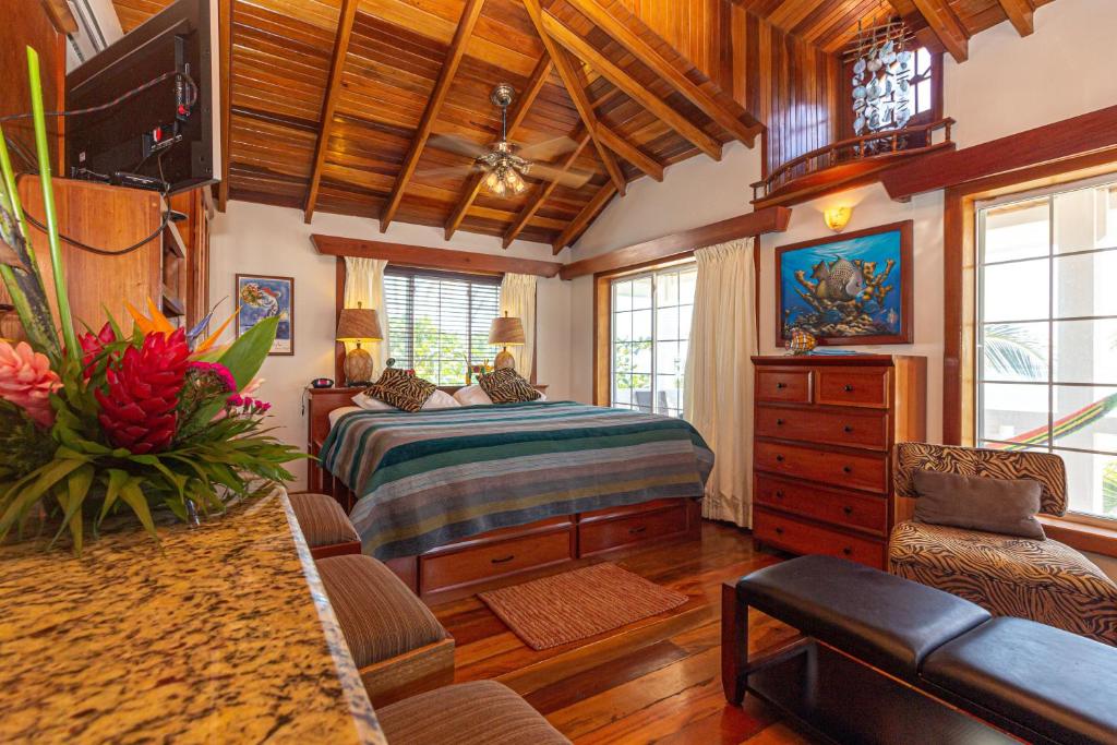 Quarto do Blue Tang Inn, para ilustrar o post sobre hotéis em Belize. O espaço é grande e possui vários detalhes e móveis em madeira. A cama é de casal e está centralizada na foto. Na cabeceira atrás dela há dois abajures e uma janela. No canto inferior esquerdo da foto podemos ver o relance de uma bancada de mármore com flores em cima. E no canto inferior direito é possível ver alguns puffs e poltronas. Atrás da poltrona, há uma cômoda de madeira, encostada na parede. O ambiente possui janelas e é bem iluminado.