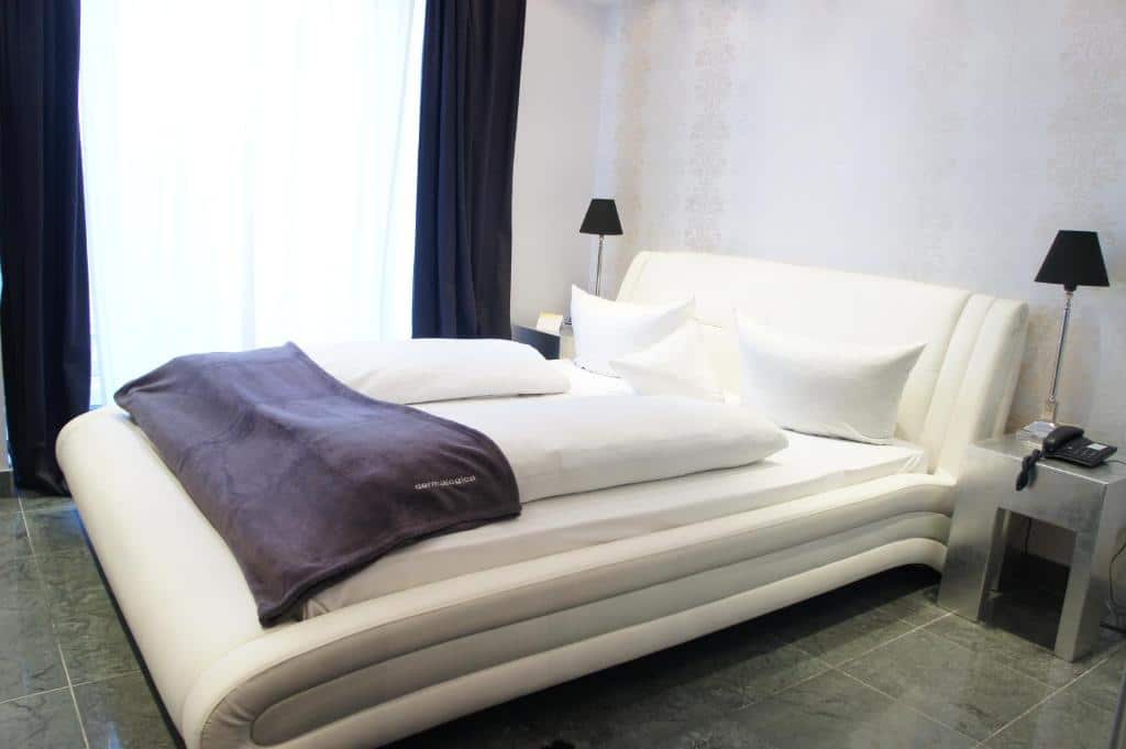 Quarto do Business Wieland Hotel com cama de casal do lado direito da imagem, com uma cômoda em cada lado com luminária. Representa hotéis em Dusseldorf.