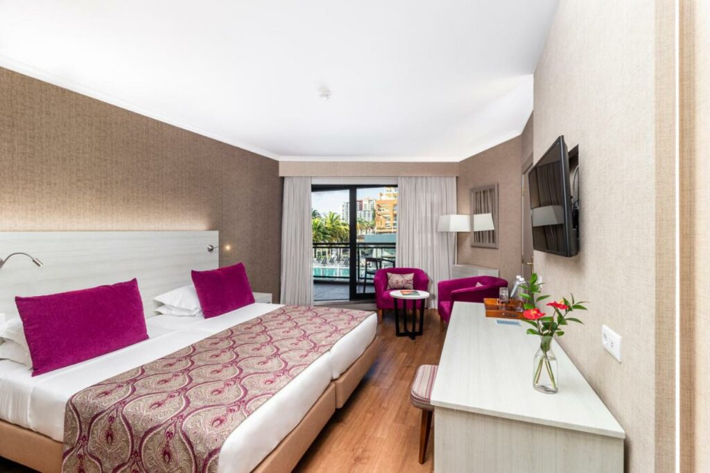 Quarto do Enotel Lido - All Inclusive com cama de casal do lado esquerdo, com uma cômoda a frente a cama e em cima da cômoda TV presa na parede e do lado esquerdo dois sofás e uma mesa redonda no meio. Representa hotéis all inclusive em Portugal.