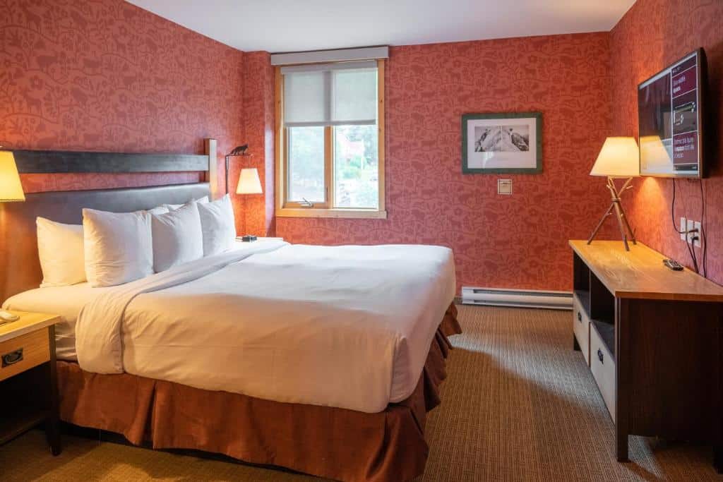 Quarto do Fox Hotel and Suites com uma cama de casal ao centro, de cada lado da cama tem uma mesinha com abajur e as paredes são vermelhas. O chão é de carpete, tem uma janela na parede e em frente a cama tem uma mesa de madeira com um abajur e uma tv na parede.