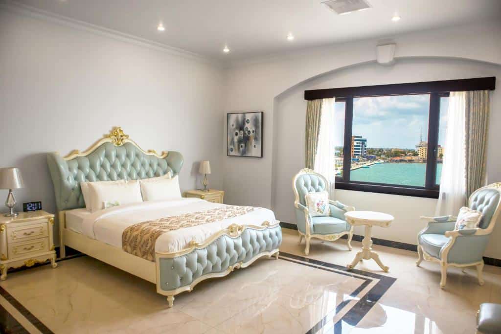 Na imagem temos a foto de um quarto no Golden Bay Belize Hotel, para ilustrar o post de hotéis em Belize. Os móveis são de estilo clássico. A cama está no lado esquerdo, centralizada no espaço. De seus dois lados há uma mesa de cabeceira com abajur em cima. Do lado direito da cama, no meio da parede do fundo, está a janela com vista para o mar, e na frente dessa janela tem duas poltronas e uma mesa de centro entre elas.