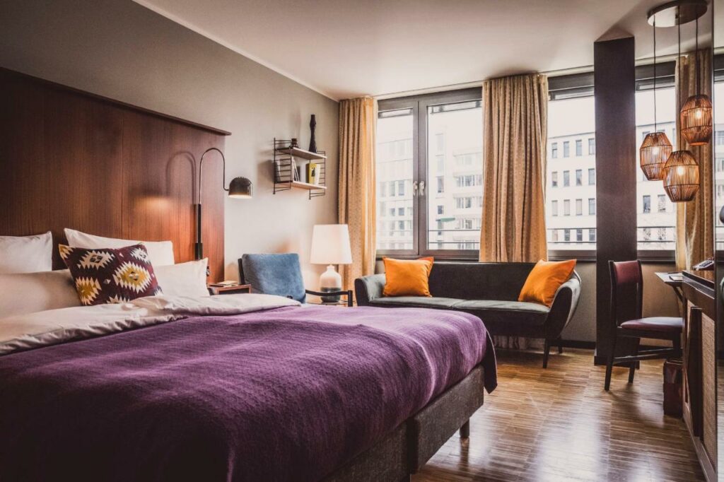 Quarto do Henri Hotel Düsseldorf Downtown com cama de casal do lado esquerdo, do lado esquerdo da cama uma cômoda, uma poltrona e um sofá. Representa hotéis em Dusseldorf.