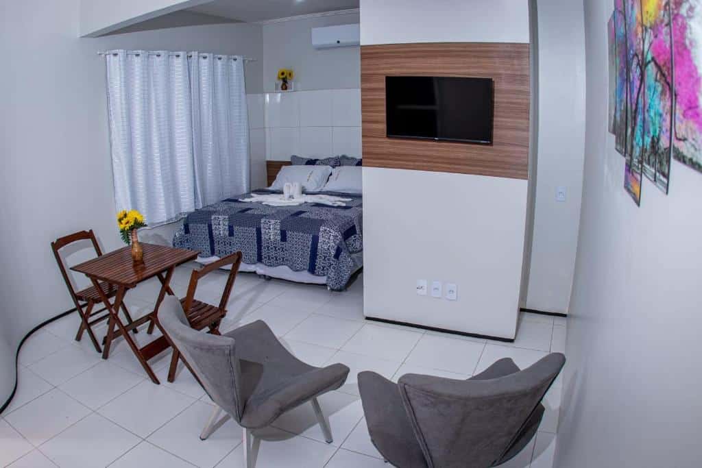 Quarto do Hotel Vila das Dunas Cumbuco com uma cama de casal no canto esquerdo da foto ao lado de uma cortina branca, em frente uma mesa quadrada e duas cadeiras de madeira e ao lado duas poltronas. Em frente as poltronas tem uma tv na parede e quadros na parede do lado.