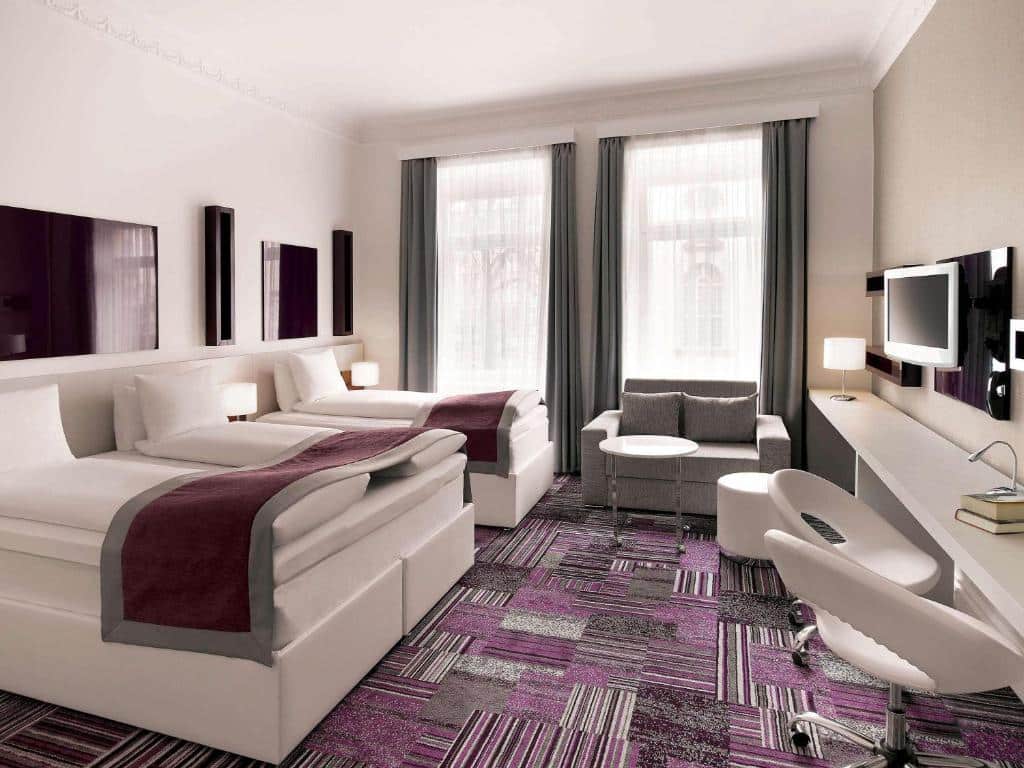 Quarto do Ibis Styles Stockholm Odenplan com cama de casal do lado esquerdo da imagem, uma cômoda ao meio com luminária e do lado esquerdo da cama de casal uma cama de solteiro. Em frente a cama de casal uma cômoda com duas cadeiras e TV presa na parede. Representa hotéis em Estocolmo.