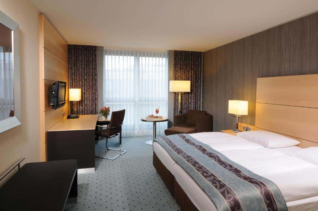 Quarto do Maritim Hotel Düsseldorf com cama de casal do lado direito da imagem, do lado esquerdo da cama uma poltrona com uma mesinha redonda e a frente uma mesa de trabalho com TV presa na parede. Representa hotéis em Dusseldorf.