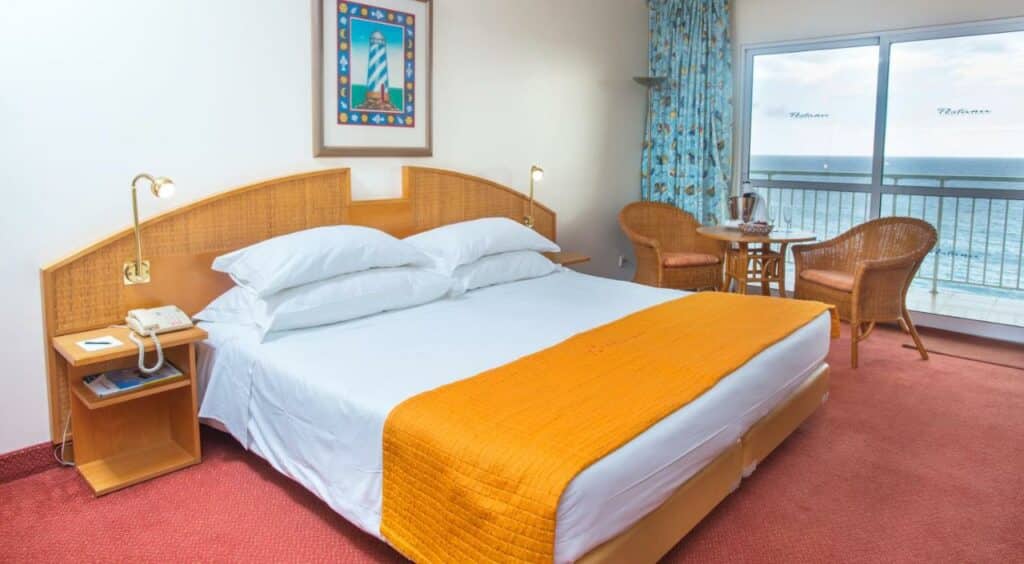 Quarto do Pestana Ocean Bay Resort com cama de casal no meio do quarto, ao lado da cama uma cômoda em cada lado, do lado direito da duas cadeiras de palhas com uma redonda no meio. Representa hotéis all inclusive em Portugal.