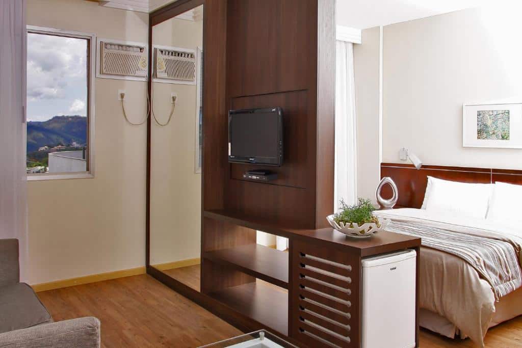 Quarto do Piemonte Hotel, representando o post sobre pousadas em Nova Lima. No lado direito, está a cama box e casal, e na sua frente há um painel de madeira que serve como divisória para a sala no outro lado do quarto. Nesse lado, no canto inferior esquerdo há um sofá, e uma TV e espelho no painel. Também há um frigobar, acoplado no painel de madeira.