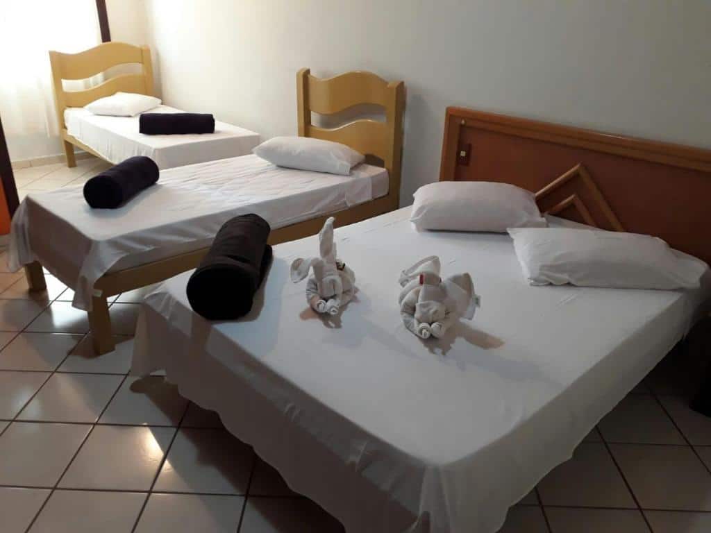 Quarto da Pousada Casa D` Guio com uma cama de casal do lado de uma cama de solteiro e mais ao fundo no canto esquerdo tem outra cama de solteiro. Todas elas com cabeceiras de madeira, com travesseiros e colcha em cima.