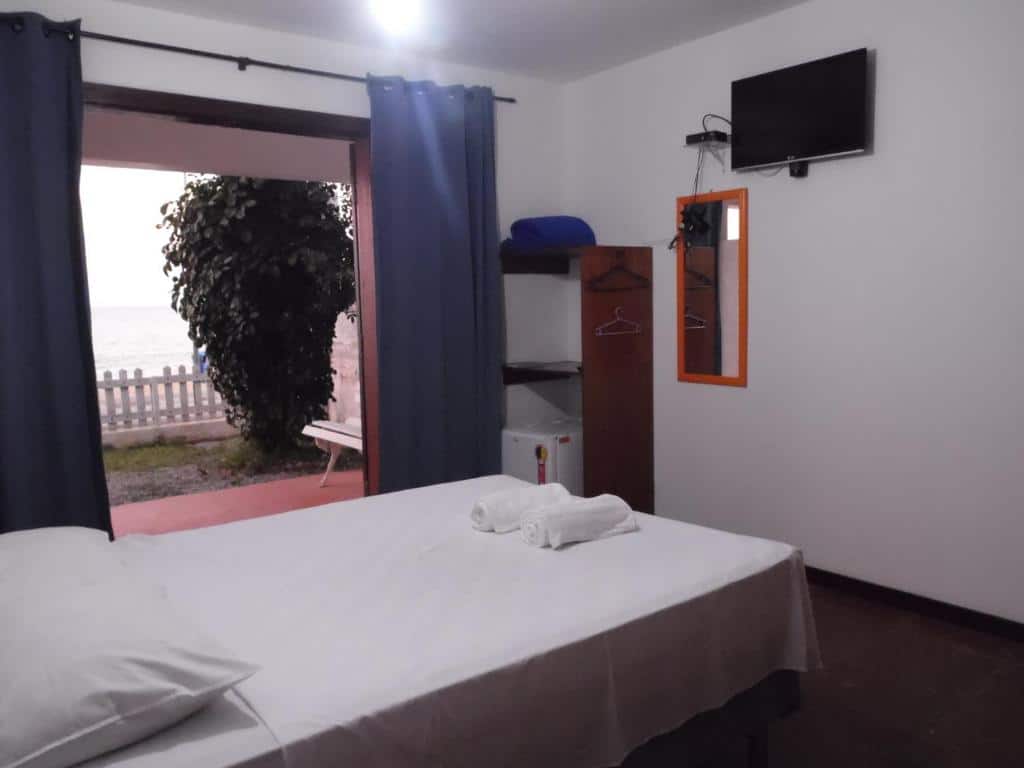 Quarto da Pousada Casa dos Sonhos com uma cama de casal ao centro, em frente uma tv, um espelho e um guarda-roupa aberto com um frigobar. Do lado tem uma porta aberta com cortinas que dá acesso a a varanda do quarto com uma árvore.