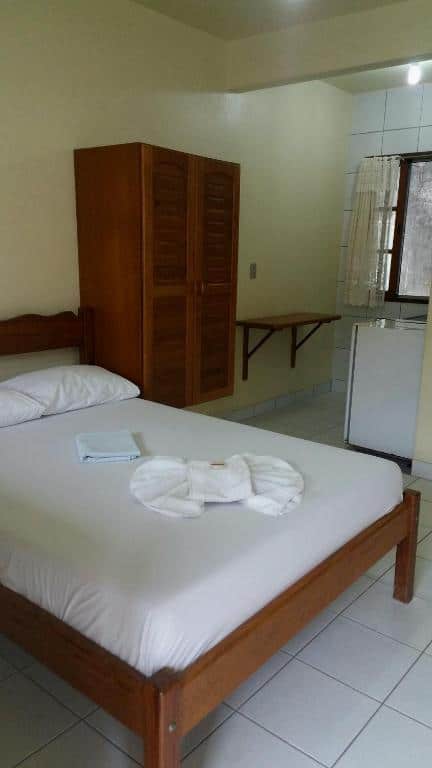 Quarto da Pousada da Nalva com uma cama de casal com dois travesseiros, ao lado da cama há um guarda-roupa de madeira, uma mesinha na parede, uma janela ao fundo e ao lado um frigobar.