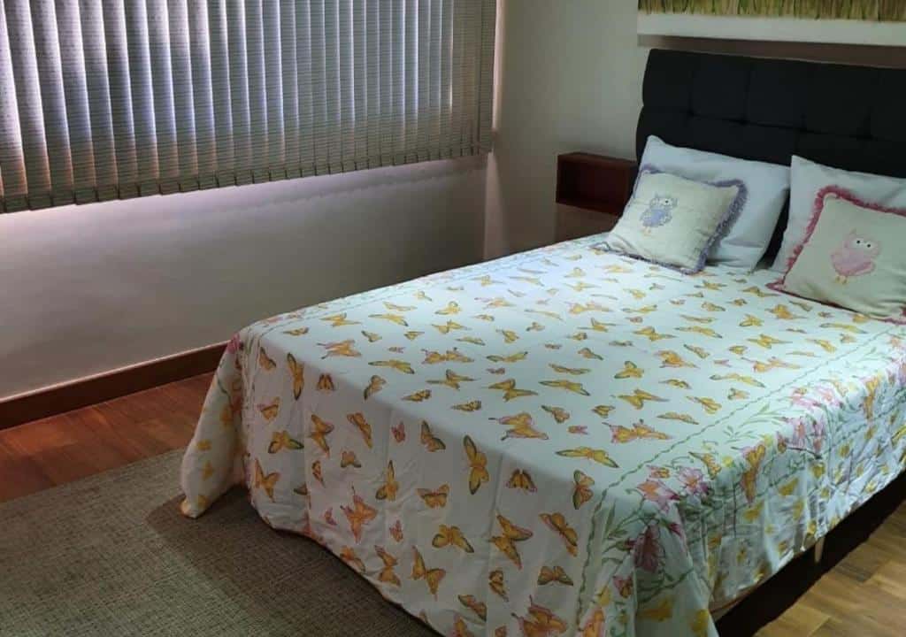 Quarto da Pousada da Vavá com uma cama de casal com travesseiros, almofadas e edredom. O quarto tem chão de madeira, um tapete no pé da cama e uma janela com persiana.
