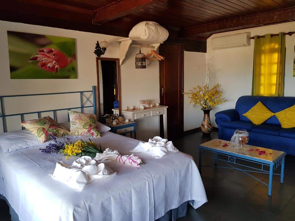 Quarto da Pousada do Marujo com uma cama de casal com almofadas e flores, em cima um quadros e ao lado duas mesinhas com vários objetos em cima e um espelho. Já ao fundo na parte direita tem um vaso grande ao lado de um sofá azul com duas almofadas e na frente uma mesinha de centro.