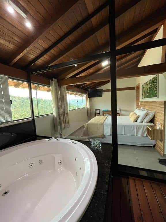 Quarto na Pousada Mar Mineiro Macacos, representando o post sobre pousadas em Nova Lima. No canto inferior esquerdo, há uma hidro, que se separa do resto da suíte por uma longa janela de vidro. Através do vidro, vemos a cama box de casal, encostada na parede direita. Na sua frente, há uma parede coberta com janelas de vidro que proporcionam uma vista para muitas árvores.