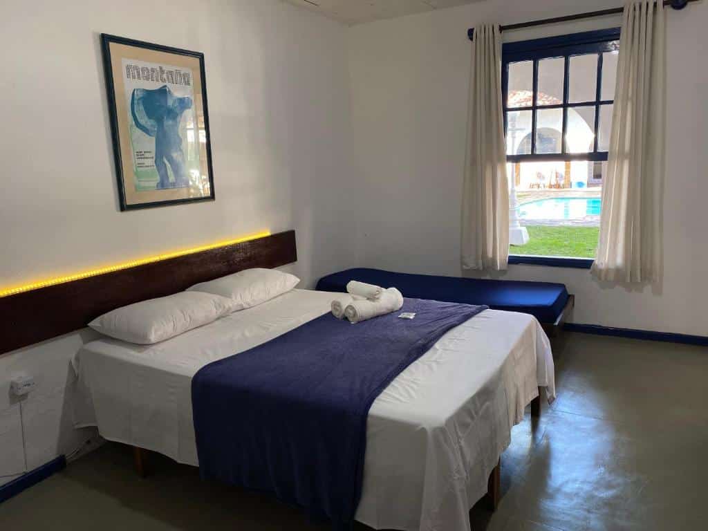 Quarto da Pousada Puerta Del Sol Rio das Ostras com uma cama de casal e uma cama de solteiro perto da janela. Além disso tem um quadro na parede e um luz em fio de led na parede também.