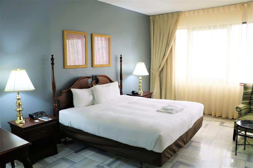 Foto do quarto do Radisson Fort George Hotel & Marina, para representar o post sobre hotéis em Belize. A cama é de casal e está no centro. Em seus dois lados há uma mesa de cabeceira com abajur e cima. Na parede atrás da cama, há dois quadros com molduras douradas presos na parede. No fundo do quarto, ao lado direito da cama, há uma janela coberta por cortinas beges. Podemos ver uma mesa redonda e uma poltrona de relance no canto inferior direito.