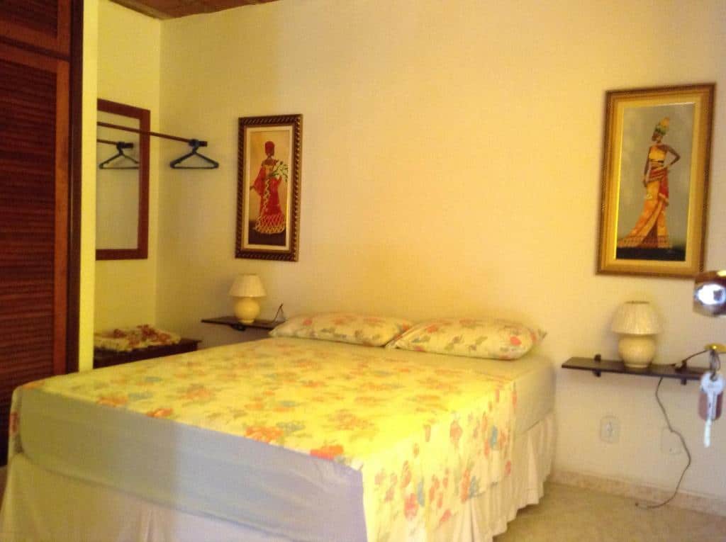 Quarto do Rey das Ostras Pousada com uma cama de casal com dois travesseiros, de cada lado da cama tem uma mesinha com abajur e dois quadros na parede. Além disso no canto esquerdo tem um espelho com uma arara de cabides e um guarda-roupa.