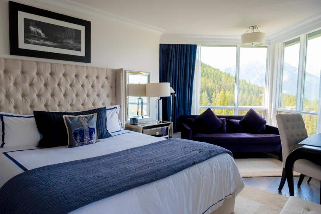 Quarto do Rimrock Resort Hotel com uma cama de casal com cabeceira almofadada, um quadro em cima e um espelho com uma mesinha e abajur ao lado. No fundo tem um sofá azul, uma poltrona estofada branca e uma parede de vidro com cortina azul.