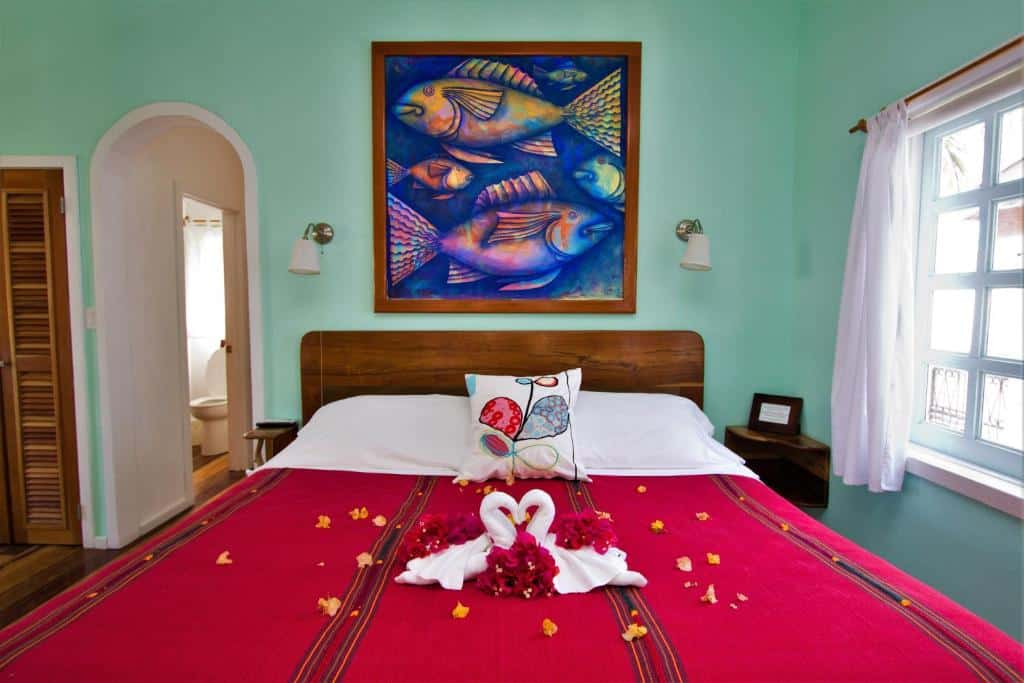 Foto de frente da cama de casal em um quarto no Sea Dreams Hotel. Há decorações românticas em cima da cama. Atrás dela, na parede, há um grande quadro com pintura de alguns peixes. No lado direito da cama há uma janela, e no esquerdo há um corredor para o banheiro.