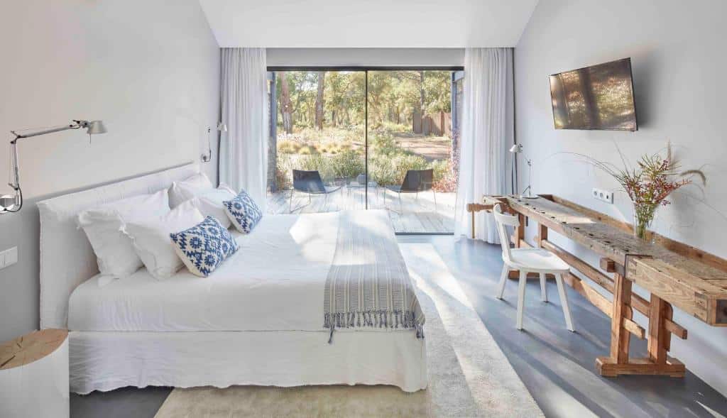 Quarto do Sublime Comporta Country Retreat & SPA, com cama de casal do lado esquerdo da imagem com uma mesa ampla em frente a cama com cadeira e em cima TV presa na parede. Representa hotéis para lua de mel em Portugal.