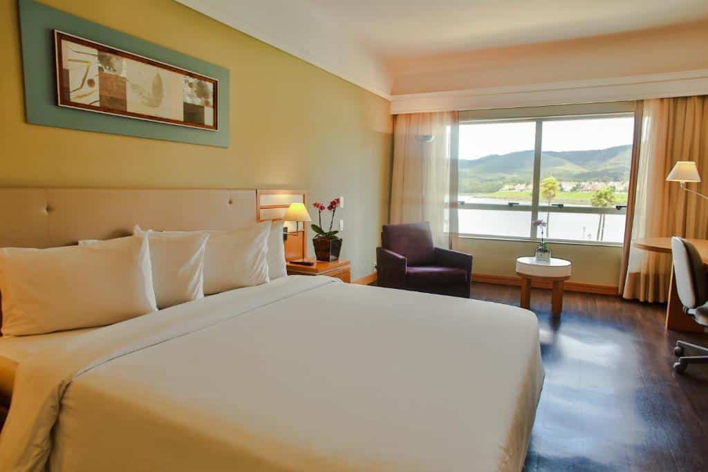 Suíte no Transamerica Lagoa dos Ingleses, representando o post sobre pousadas em Nova Lima. A cama é larga, box e de casal, e na foto ela está no canto inferior esquerdo. Do seu lado direito, há uma mesa de cabeceira com abajur e um vaso com flores. Mais a frente, há uma poltrona do lado de uma pequena mesa circular. No canto direito, vemos parcialmente uma cadeira giratória e uma escrivaninha com outro abajur. No fundo do quarto, estão as janelas com cortinas beges.