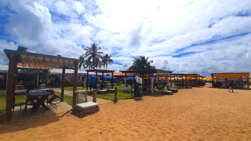 Areia da praia do Gunga, com mesas e áreas cobertas com bancos e rede de vários quiosques